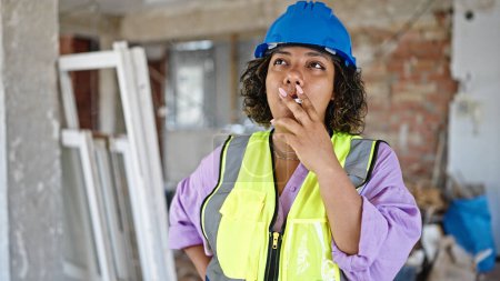 Foto de Joven hermosa mujer latina constructor fumar cigarrillo con cara seria en el sitio de construcción - Imagen libre de derechos