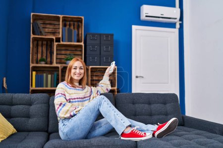Téléchargez les photos : Jeune femme caucasienne utilisant la machine de climatisation assis sur le canapé à la maison - en image libre de droit