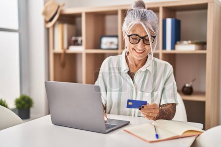 Photo for Middle age grey-haired woman using laptop and credit card sitting on table at home - Royalty Free Image