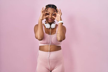 Foto de Mujer afroamericana con trenzas que usan ropa deportiva y auriculares tratando de abrir los ojos con los dedos, soñolienta y cansada de fatiga matutina - Imagen libre de derechos