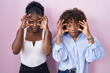 Téléchargez les photos : Deux femmes africaines debout sur fond rose essayant d'ouvrir les yeux avec les doigts, somnolentes et fatiguées pour la fatigue matinale - en image libre de droit
