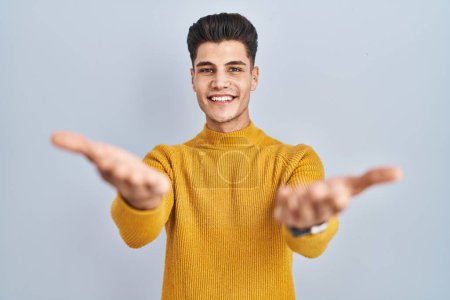 Téléchargez les photos : Jeune homme hispanique debout sur fond bleu souriant joyeuse offrant des mains donnant assistance et acceptation. - en image libre de droit