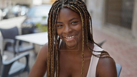 Foto de Mujer afroamericana sonriendo confiada sentada en la mesa en la terraza de la cafetería - Imagen libre de derechos
