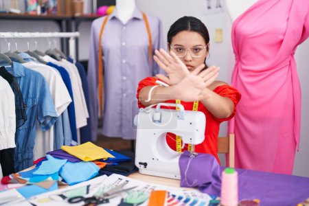 Photo for Hispanic young woman dressmaker designer using sewing machine rejection expression crossing arms and palms doing negative sign, angry face - Royalty Free Image