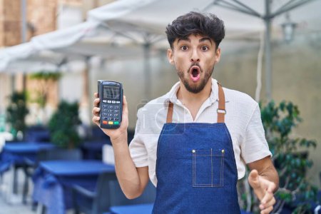Photo for Arab man with beard wearing waiter apron at restaurant terrace holding dataphone scared and amazed with open mouth for surprise, disbelief face - Royalty Free Image