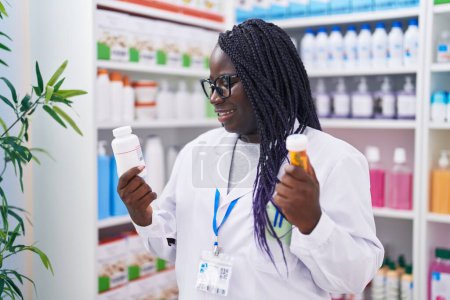 Téléchargez les photos : Afro-américaine femme pharmacien souriant confiant tenant bouteilles pilules à la pharmacie - en image libre de droit