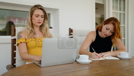 Foto de Dos mujeres usando la escritura del ordenador portátil en el cuaderno sentado en la mesa en la terraza del hogar - Imagen libre de derechos
