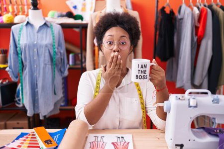 Photo for Beautiful african woman with curly hair dressmaker designer drinking from i am the boss cup covering mouth with hand, shocked and afraid for mistake. surprised expression - Royalty Free Image