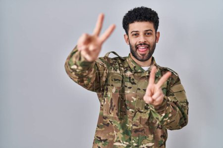 Foto de Hombre árabe vistiendo uniforme camuflaje del ejército sonriendo con la lengua hacia fuera mostrando los dedos de ambas manos haciendo signo de victoria. número dos. - Imagen libre de derechos