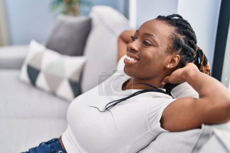 Téléchargez les photos : Femme afro-américaine détendue avec les mains sur la tête assise sur le canapé à la maison - en image libre de droit