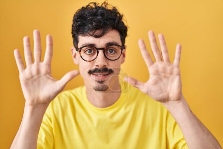 Photo for Hispanic man wearing glasses standing over yellow background showing and pointing up with fingers number ten while smiling confident and happy. - Royalty Free Image