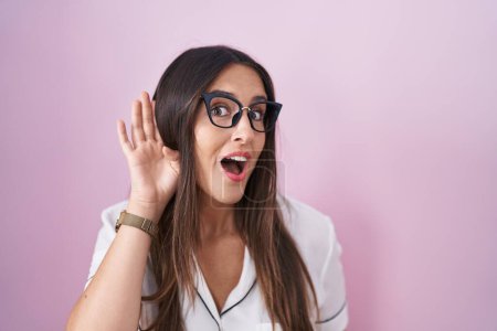 Téléchargez les photos : Jeune femme brune portant des lunettes debout sur fond rose souriant avec la main sur l'oreille en écoutant une rumeur ou des commérages. concept de surdité. - en image libre de droit