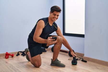 Téléchargez les photos : Jeune homme hispanique utilisant kettlebell et smartphone formation au centre sportif - en image libre de droit