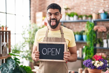 Photo for Hispanic man with beard working at florist holding open sign sticking tongue out happy with funny expression. - Royalty Free Image