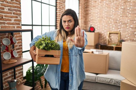 Téléchargez les photos : Jeune femme hispanique déménageant dans une nouvelle maison tenant des plantes avec la main ouverte faisant stop signe avec expression sérieuse et confiante, geste de défense - en image libre de droit
