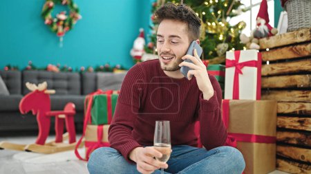 Photo for Young hispanic man talking on smartphone drinking champagne celebrating christmas at home - Royalty Free Image