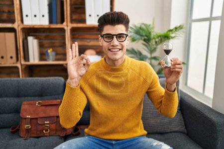 Foto de Joven hombre hispano sosteniendo reloj de arena haciendo señal bien con los dedos, sonriendo gesto amistoso excelente símbolo - Imagen libre de derechos