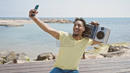 Foto de Hombre afroamericano sosteniendo boombox hacer selfie por teléfono inteligente en la playa - Imagen libre de derechos