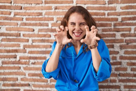 Téléchargez les photos : Belle femme brune debout sur un mur de briques souriant drôle faisant griffe geste comme chat, expression agressive et sexy - en image libre de droit