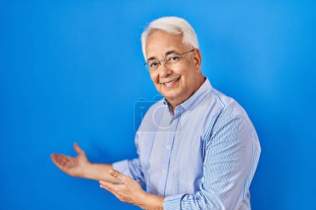 Photo for Hispanic senior man wearing glasses inviting to enter smiling natural with open hand - Royalty Free Image
