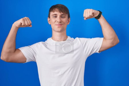 Foto de Hombre rubio caucásico de pie sobre fondo azul mostrando los músculos de los brazos sonriendo orgulloso. concepto de fitness. - Imagen libre de derechos
