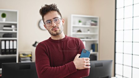 Téléchargez les photos : Jeune homme hispanique travailleur d'affaires debout avec les bras croisés geste tenant smartphone au bureau - en image libre de droit