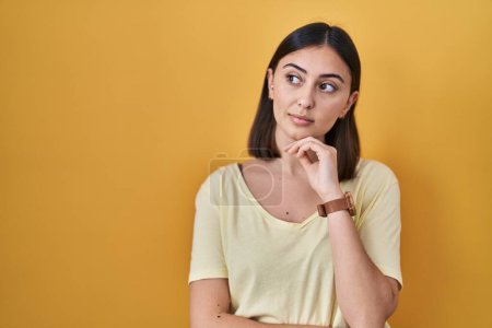 Photo for Hispanic girl wearing casual t shirt over yellow background with hand on chin thinking about question, pensive expression. smiling with thoughtful face. doubt concept. - Royalty Free Image