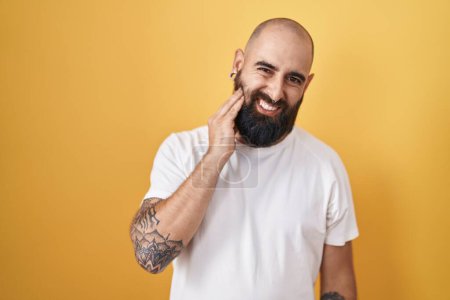 Foto de Joven hombre hispano con barba y tatuajes de pie sobre fondo amarillo tocando boca con mano con expresión dolorosa debido a dolor de muelas o enfermedad dental en los dientes. dentista - Imagen libre de derechos