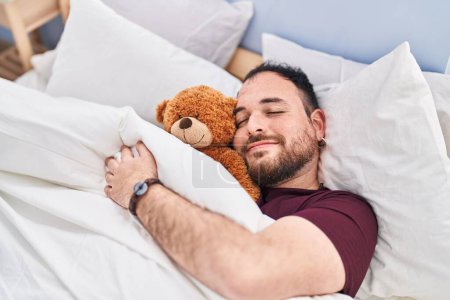 Photo for Young hispanic man hugging teddy bear lying on bed sleeping at bedroom - Royalty Free Image