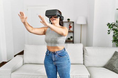 Téléchargez les photos : Jeune belle femme hispanique jouer à un jeu vidéo en utilisant des lunettes de réalité virtuelle à la maison - en image libre de droit
