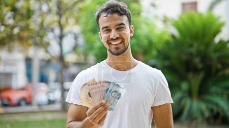 Photo for Young hispanic man smiling confident holding mexican pesos at park - Royalty Free Image