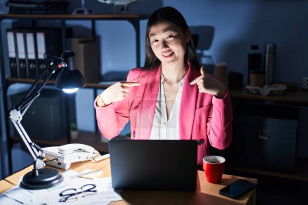 Foto de Mujer joven china que trabaja en la oficina por la noche mirando confiado con sonrisa en la cara, señalándose con los dedos orgullosos y felices. - Imagen libre de derechos