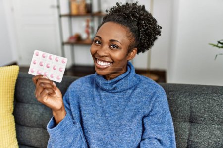 Téléchargez les photos : Femme afro-américaine tenant des pilules assis sur le canapé à la maison - en image libre de droit