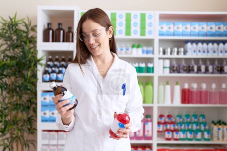 Téléchargez les photos : Jeune belle pharmacienne souriante confiante tenant des bouteilles de médicaments à la pharmacie - en image libre de droit