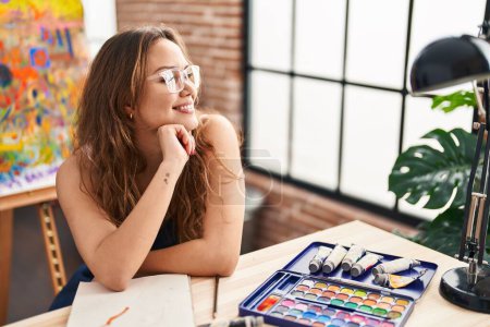 Téléchargez les photos : Jeune belle hispanique femme artiste souriant confiant assis sur la table à l'atelier d'art - en image libre de droit