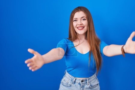 Photo for Redhead woman standing over blue background looking at the camera smiling with open arms for hug. cheerful expression embracing happiness. - Royalty Free Image