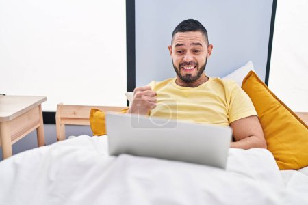 Foto de Hombre hispano usando portátil en la cama celebrando el logro con sonrisa feliz y expresión ganadora con la mano levantada - Imagen libre de derechos