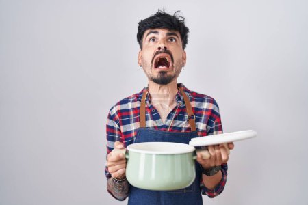 Photo for Young hispanic man with beard wearing apron holding cooking pot angry and mad screaming frustrated and furious, shouting with anger looking up. - Royalty Free Image
