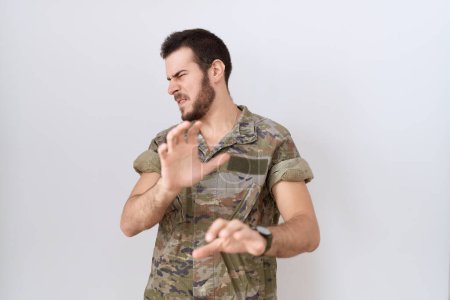 Foto de Joven hombre hispano con uniforme camuflaje del ejército expresión repugnante, disgustado y temeroso haciendo cara de asco porque la reacción de aversión. con las manos levantadas - Imagen libre de derechos