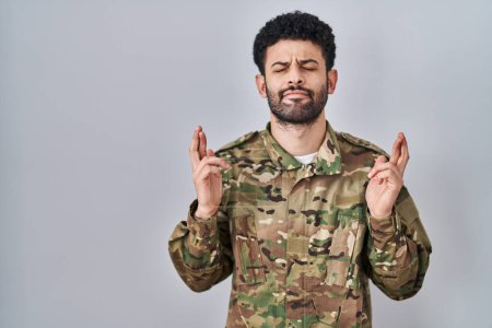 Téléchargez les photos : Homme arabe portant uniforme de camouflage militaire geste doigt croisé souriant avec espoir et les yeux fermés. chance et concept superstitieux. - en image libre de droit