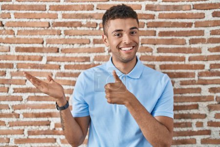 Téléchargez les photos : Jeune homme brésilien debout sur un mur de briques montrant la main de la paume et faisant un geste correct avec les pouces levés, souriant heureux et joyeux - en image libre de droit