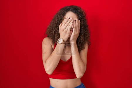 Téléchargez les photos : Femme hispanique avec les cheveux bouclés debout sur fond rouge frottant les yeux pour la fatigue et les maux de tête, somnolent et l'expression fatiguée. problème de vision - en image libre de droit