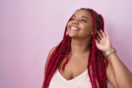 Téléchargez les photos : Femme afro-américaine avec les cheveux tressés debout sur fond rose souriant cheveux touchants confiants avec geste de la main vers le haut, posant attrayant et à la mode - en image libre de droit