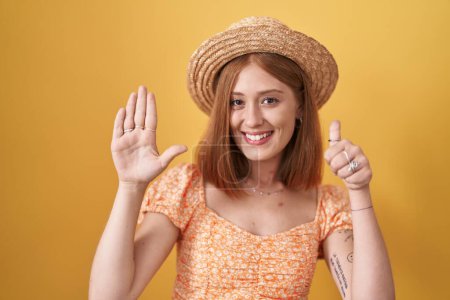 Foto de Joven pelirroja de pie sobre fondo amarillo con sombrero de verano mostrando y señalando hacia arriba con los dedos número seis mientras sonríe confiado y feliz. - Imagen libre de derechos