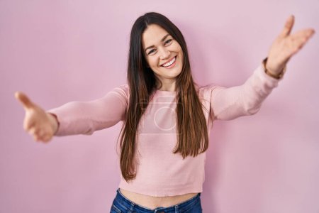 Téléchargez les photos : Young brunette woman standing over pink background looking at the camera smiling with open arms for hug. cheerful expression embracing happiness. - en image libre de droit