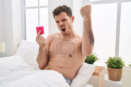 Foto de Joven hombre hispano sosteniendo condón en la cama molesto y frustrado gritando con ira, gritando loco de ira y mano levantada - Imagen libre de derechos
