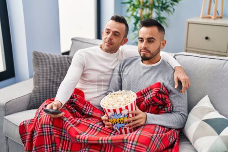 Téléchargez les photos : Couple homosexuel manger du maïs soufflé regarder la télévision détendue avec une expression sérieuse sur le visage. simple et naturel regardant la caméra. - en image libre de droit