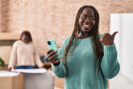 Téléchargez les photos : Femme africaine utilisant smartphone à la nouvelle maison pointant pouce vers le côté souriant heureux avec bouche ouverte - en image libre de droit