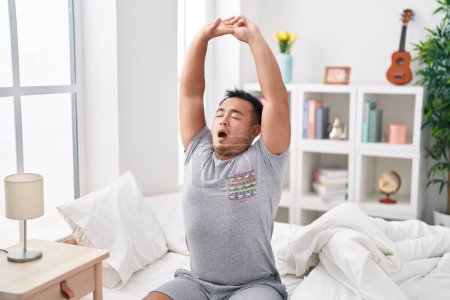 Photo for Young chinese man waking up stretching arms yawning at bedroom - Royalty Free Image