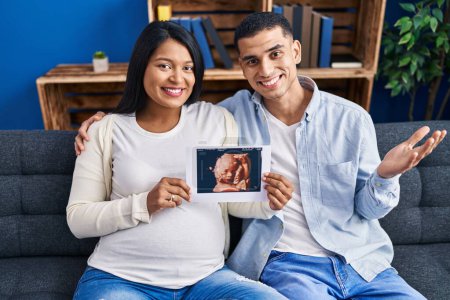 Photo for Young hispanic couple expecting a baby sitting on the sofa showing baby ultrasound celebrating achievement with happy smile and winner expression with raised hand - Royalty Free Image